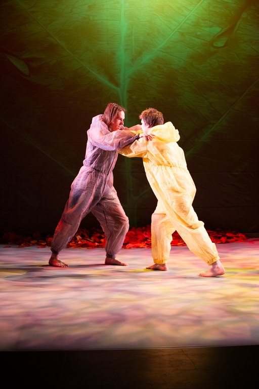 I bilden syns två medverkande. De lutar sig mot varandra ansikte mot ansikte, med stöd av sina händer. Den ena medverkande bär en grå overall, den andra en gul.