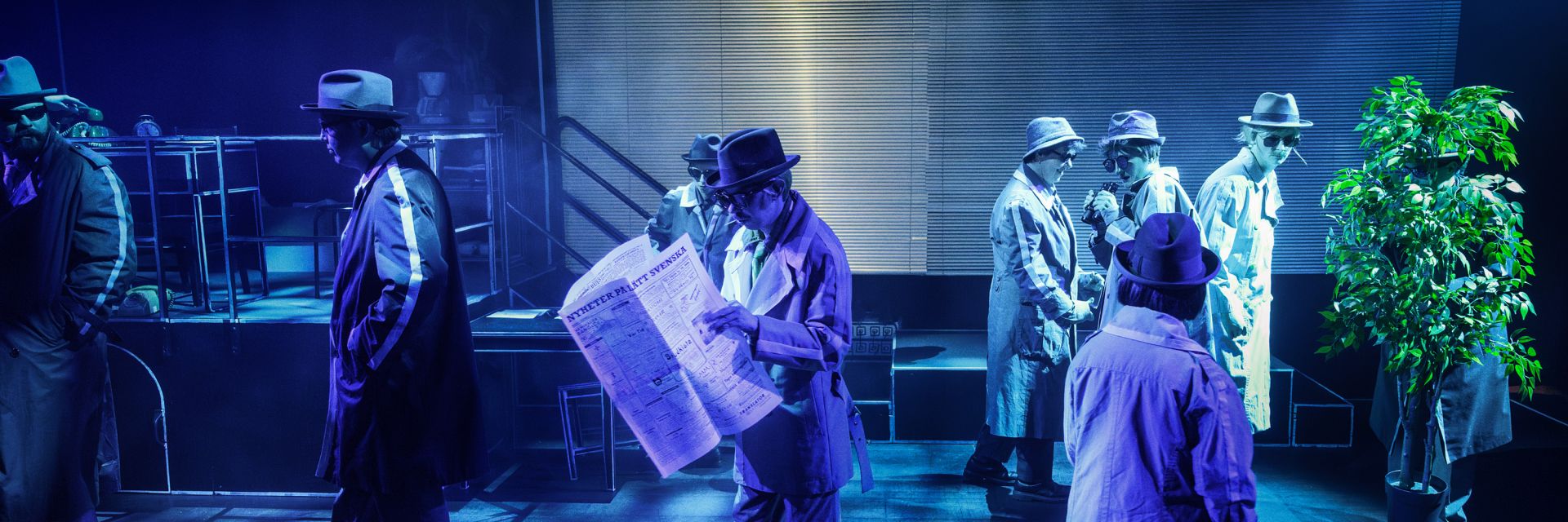 En scen fylld av skådespelare i trenchcoats, hattar och solglasögon, upplysta av blått och lila ljus som skapar en mystisk film noir-stämning. I förgrunden läser en person en stor tidning med rubriken ’NYHETER PÅ RIKT SVENSKA’. Till vänster står en annan gestalt med händerna i fickorna, medan flera figurer i bakgrunden rör sig i olika riktningar som om de håller på med hemliga uppdrag. Två personer står tätt ihop och undersöker något i händerna, medan en tredje figur längre bak tuggar på ett sugrör eller cigarettmunstycke. Till höger försöker en skådespelare gömma sig bakom en stor krukväxt, vilket förstärker scenens humoristiska och parodiska spionkänsla.