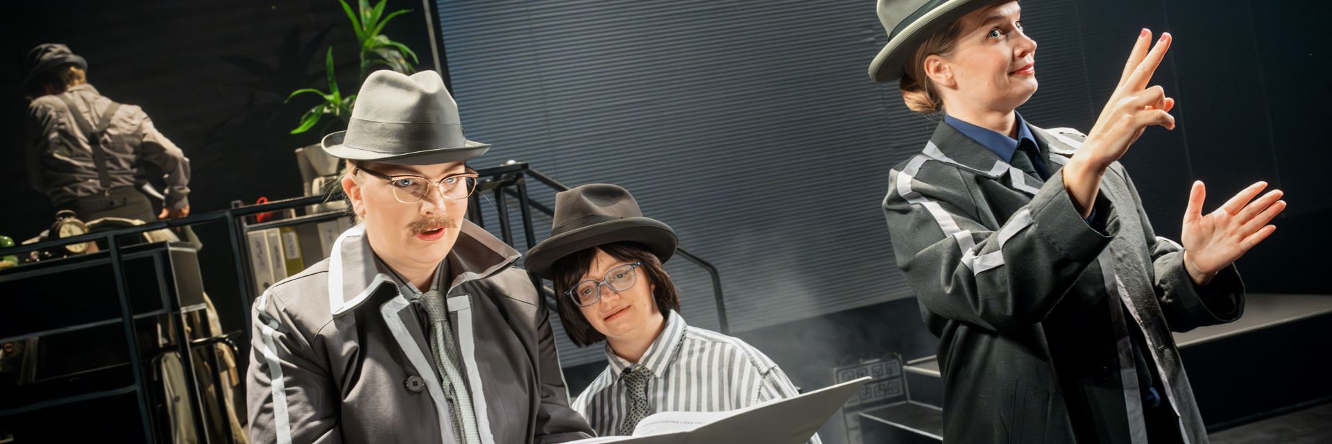Tre skådespelare står i ett upplyst scenrum iförda grå kostymer, slipsar och bredbrättade hattar i en stil som påminner om klassiska deckare. Personen längst till vänster står i profil och läser i ett dokument med koncentrerad min, iförd runda glasögon och en markerad mustasch. Bredvid hen ler en annan skådespelare med mörk peruk, hatt och stora glasögon, också med dokument i handen. Till höger står en tredje person, mer framlyft i ljuset, och gestikulerar uttrycksfullt med händerna som om hon förklarar något viktigt. I bakgrunden syns suddiga kontorsmöbler och pappersmappar, vilket skapar en miljö som liknar ett gammaldags detektivkontor.