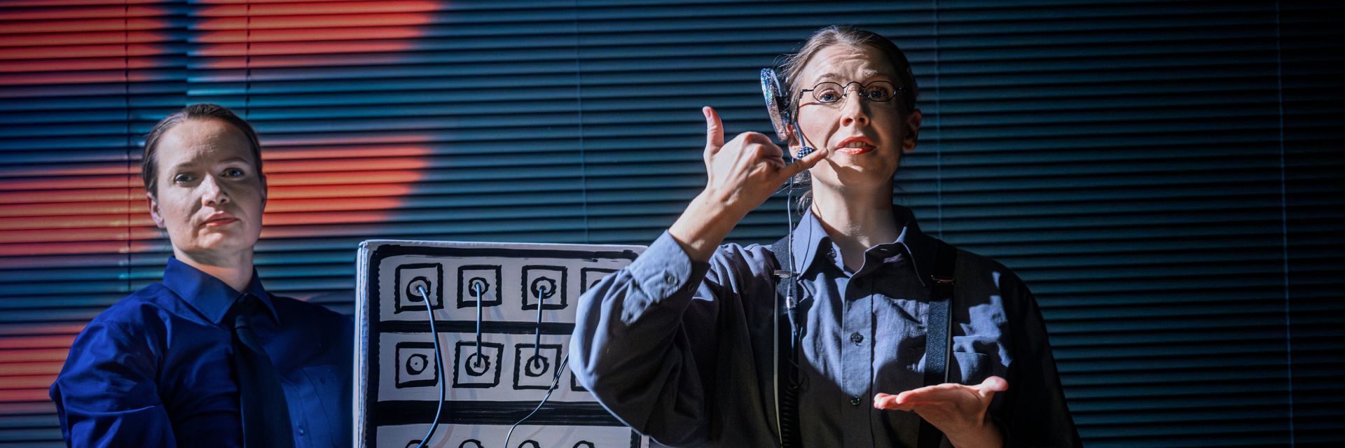 Två skådespelare står framför en bakgrund av horisontella persienner, upplysta av starka färgskiftande scenljus. Personen till höger bär glasögon, headset och grå skjorta med hängslen, och gestikulerar med handen i en tydlig ’ringa’-symbol medan hen håller den andra handen öppen framför sig som om hen instruerar eller demonstrerar något. Bredvid står en skådespelare i mörkblå skjorta och slips, med allvarlig min och blicken riktad rakt fram. I mitten syns en stor rekvisita som liknar ett gammaldags telefonväxelblock med handritade symboler och sladdar som hänger ut. Ljuset och poserna skapar en känsla av både humor och precision i en stiliserad kontorsmiljö.