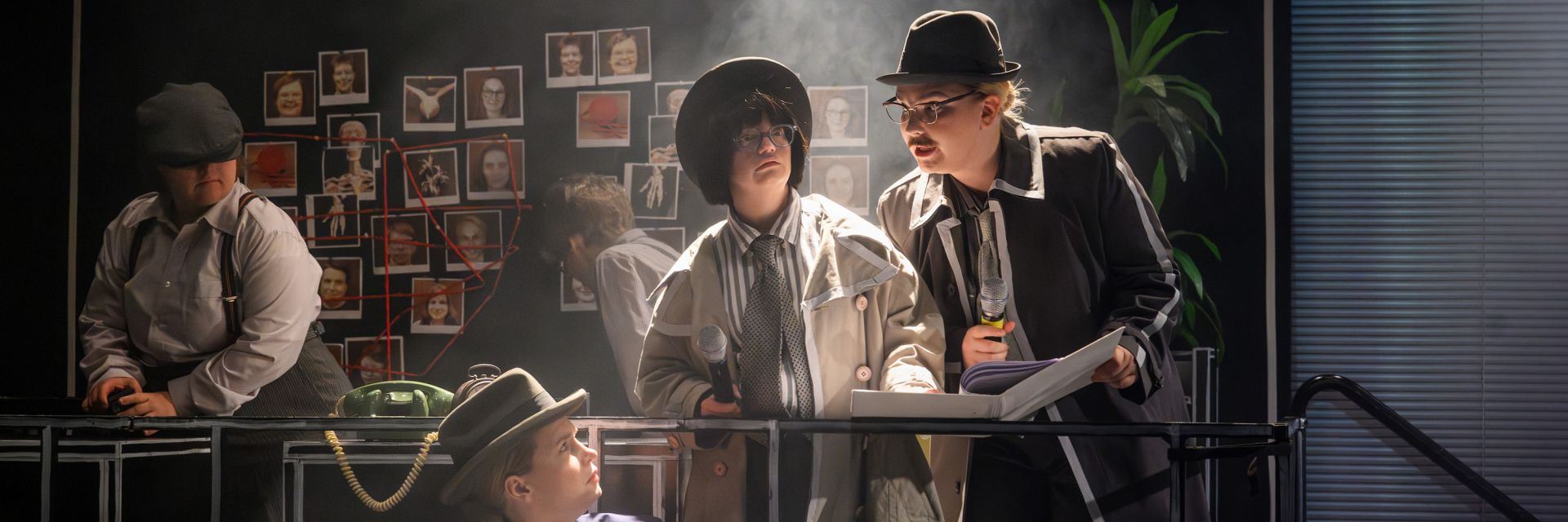 En grupp skådespelare står i en scen som liknar ett detektivkontor. I bakgrunden syns en vägg täckt av fotografier och bilder sammanbundna med röda trådar, som i en klassisk brottsutredning. Två skådespelare i förgrunden, båda i trenchcoat, hatt och slips, håller mikrofoner och studerar ett öppet block; den ena tittar koncentrerat ner i boken medan den andra lutar sig fram för att läsa. Till vänster står en person i skjorta, hängslen och keps och arbetar vid ett bord med gamla telefoner. En annan skådespelare sitter längre fram och tittar upp mot sina kollegor. Atmosfären är dimmig och belyst från sidan, vilket förstärker känslan av ett hemligt och intensivt detektivarbete.