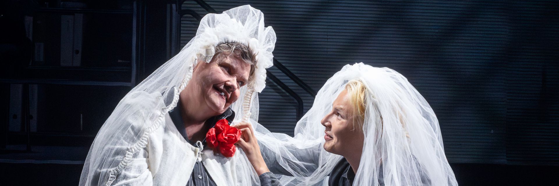 Två skådespelare står nära varandra på scen, båda iklädda genomskinliga, vita bröllopsslöjor som faller över deras ansikten och axlar. Personen till vänster ler varmt och lutar huvudet mjukt åt sidan medan en röd tygros sitter fäst vid bröstet på deras ljusa klädsel. Personen till höger, med ljust hår synligt under slöjan, håller varsamt den andres axel och ser på hen med en öm och tröstande blick. Bakgrunden är mörk och suddig, vilket framhäver den intima och känslosamma stunden mellan dem.